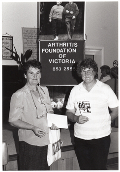 B&W photo of two women standing in front of a small stage. One woman is holding some paper and a plastic bag in one hand, while the other is holding two "Arthritis" booklets. Behind them, there is a vertical "Arthritis Foundation of Victoria" banner. Above the banner, there is a large photo poster of two people walking, and on the panel below, a large photo featuring the Joint Action Group (partially obscured). In the background, there are two framed pictures on the wall, and a potted plant on a stand next to a piano (partially obscured). To the right of the frame, there are two people standing, facing a servery.
