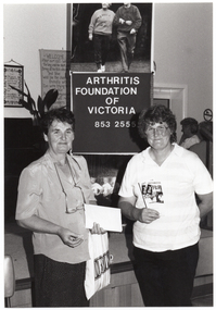 B&W photo of two women standing in front of a small stage. One woman is holding some paper and a plastic bag in one hand, while the other is holding two "Arthritis" booklets. Behind them, there is a vertical "Arthritis Foundation of Victoria" banner. Above the banner, there is a large photo poster of two people walking, and on the panel below, a large photo featuring the Joint Action Group (partially obscured). In the background, there are two framed pictures on the wall, and a potted plant on a stand next to a piano (partially obscured). To the right of the frame, there are two people standing, facing a servery.
