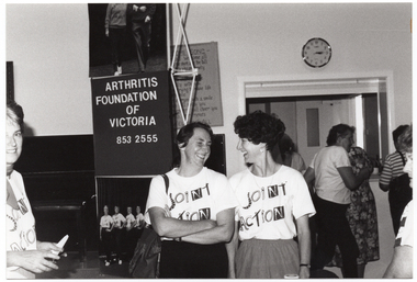 B&W photo of a small group of women in a hall. Two women (in the centre of the frame) are standing next to one another, laughing. They are both wearing t-shirts with the "Joint Action" logo. Another woman is standing to the left of the frame (partially obscured). She is looking directly at the camera. On the stage, there is a vertical "Arthritis Foundation of Victoria" banner with large photo of two people walking on the top panel, and a large photo featuring the Joint Action Group (partially obscured) on the bottom panel. Behind the banner, there is a tapestry hanging on the wall. In the background, there are a few people standing near an open servery.