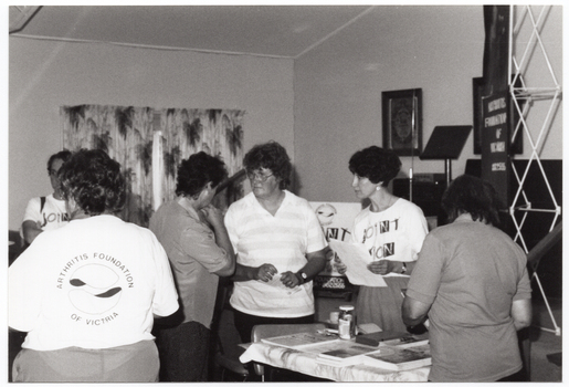 B&W photo of a small group of women in the middle of a discussion. They are standing near a table with some books and brochures on top. One woman is wearing an 'Arthritis Foundation of Victoria' t-shirt, while another woman is wearing a 'Joint Action' t-shirt. There are a few other women standing nearby.