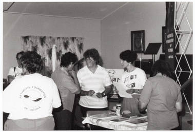 B&W photo of a small group of women in the middle of a discussion. They are standing near a table with some books and brochures on top. One woman is wearing an 'Arthritis Foundation of Victoria' t-shirt, while another woman is wearing a 'Joint Action' t-shirt. There are a few other women standing nearby.