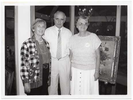 B&W photo of three people standing in front of a framed painting on a stand or easel. One woman is wearing a badge or sticker on her top with the text "ARTHRITIS WEEK". Another framed painting, on the floor and leaning against something, is evident to the left of the frame. The reflection of a chandelier light fitting is evident in the windows behind them.