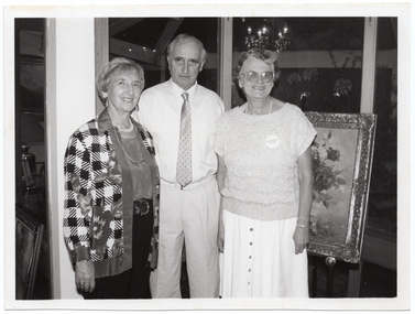 B&W photo of three people standing in front of a framed painting on a stand or easel. One woman is wearing a badge or sticker on her top with the text "ARTHRITIS WEEK". Another framed painting, on the floor and leaning against something, is evident to the left of the frame. The reflection of a chandelier light fitting is evident in the windows behind them.