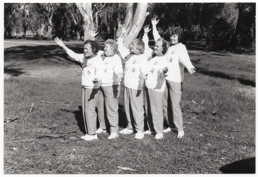 B&W photo of a five women posing for a photo in a park. They are standing close to one another in a line, at a 45 degree angle, with both arms outstretched. They are each wearing a sweatshirt printed with the "Joint Action" logo.  There are several trees in the background, with one very large tree directly behind them.