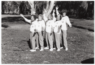 B&W photo of a five women posing for a photo in a park. They are standing close to one another in a line, at a 45 degree angle, with both arms outstretched. They are each wearing a sweatshirt printed with the "Joint Action" logo.  There are several trees in the background, with one very large tree directly behind them.