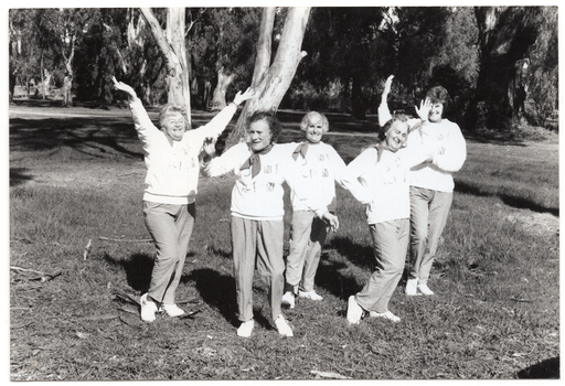 B&W photo of a five women posing for a photo in a park. They are standing close to one another in a line, at a 45 degree angle. Each woman is striking an individual pose. They are each wearing a sweatshirt printed with the "Joint Action" logo.  There are several trees in the background, with one very large tree directly behind them.