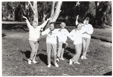B&W photo of a five women posing for a photo in a park. They are standing close to one another in a line, at a 45 degree angle. Each woman is striking an individual pose. They are each wearing a sweatshirt printed with the "Joint Action" logo.  There are several trees in the background, with one very large tree directly behind them.