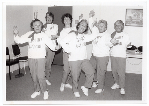 B&W photo of a small group of women posing for a photo near a doorway in the corner of a room. They are each wearing sweatshirts with the logo, "Joint Action" and track suit pants. Each woman is striking an individual pose. Behind them, there are some chairs against one wall, a closed door, a table against another wall with some miscellaneous items on top, and a painting on the wall above the table.