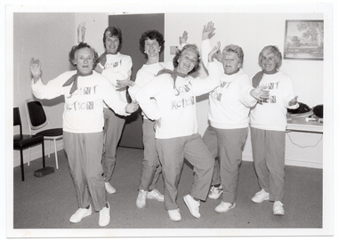 B&W photo of a small group of women posing for a photo near a doorway in the corner of a room. They are each wearing sweatshirts with the logo, "Joint Action" and track suit pants. Each woman is striking an individual pose. Behind them, there are some chairs against one wall, a closed door, a table against another wall with some miscellaneous items on top, and a painting on the wall above the table.