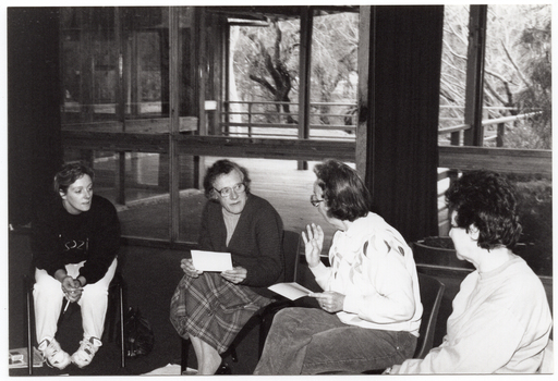 B&W photo of four women, sitting on chairs in a semi-circle. One woman is speaking while the other three women are facing her.