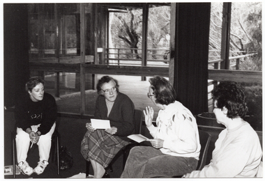 B&W photo of four women, sitting on chairs in a semi-circle. One woman is speaking while the other three women are facing her.