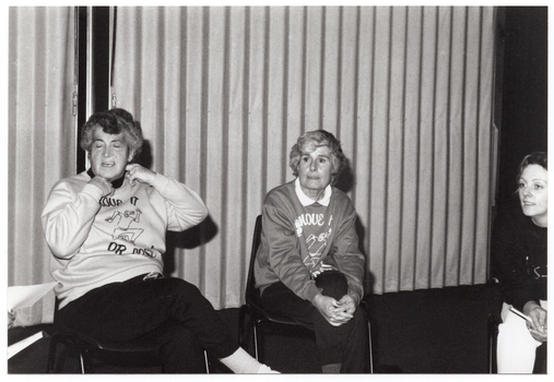 B&W photo of three women (one partially obscured), sitting on chairs in a large room. Two of the women are wearing 'Move it or Lose it' sweatshits. Behind them, there is a set of large glass sliding doors covered by closed curtains.