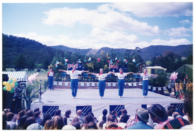 COL photo of a group of women presenting an exercise demonstration on a large outdoor stage. Standing in the rear corner of the stage is another woman.  At the back of the stage is a dais, with chairs laid out in a row. The dais has been decorated with red, white, and blue balloons. To the left of the frame, there is a blue and white striped market gazebo. In front of it, there is a camera operator standing off stage, filming the performers on the stage. There is a big bunch of yellow and green balloons just behind him. The front of the stage is covered with a long banner containing multiple sections with the VicHealth Foundation text and logo. Members of the audience, who sitting on the grass in front of the high stage, are evident in the foreground and have been Photographed from behind. A mountain range covered in trees is evident in the background. The photo has been taken from the front, directly facing the stage.