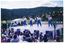 COL photo of a group of women presenting an exercise demonstration on a large outdoor stage. Standing in the rear corner of the stage is another woman. At the back of the stage is a dais, with chair laid out in a row. The dais has been decorated with red, white, and blue balloons. To the left of the stage, there is a blue and white striped market gazebo and a big bunch of yellow and green balloons tied to a stack of PA speakers. The front of the stage is covered with a long banner containing multiple sections with the VicHealth Foundation text and logo. Members of the audience, who sitting on the grass in front of the high stage, are evident in the foreground. They have been Photographed from behind, slightly to one side at a 45 degree angle. A mountain range covered in trees is evident in the background.