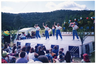 COL photo of a group of women presenting an exercise demonstration on a large outdoor stage. Standing in the rear corner of the stage is another woman. At the back of the stage is a dais, with chair laid out in a row. The dais has been decorated with red, white, and blue balloons. To the left of the stage, there is a blue and white striped market gazebo and a big bunch of yellow and green balloons tied to a stack of PA speakers. The front of the stage is covered with a long banner containing multiple sections with the VicHealth Foundation text and logo. Members of the audience, who sitting on the grass in front of the high stage, are evident in the foreground. They have been Photographed from behind, slightly to one side at a 45 degree angle. A mountain range covered in trees is evident in the background.