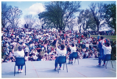 COL photo of a group of women presenting an exercise demonstration on a large outdoor stage. One woman is standing in the front corner of the stage, while the other four women are sitting on chairs arranged in a staggered formation on the stage. The performers have been Photographed from behind, and are therefore in the foreground of the image. In the background, there is a large audience sitting on a grassy hill, which slopes down towards the stage and  performers. There are several large trees behind the audience. In the distance, to the right of the frame, there is a crowd of people standing behind a string of bunting tied to some poles at waist height.