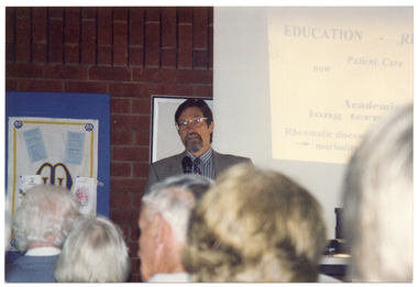 COL photo of a man standing in front of a large slide projector screen. The text on the slide being projected onto the screen (partially obscured) is: "EDUCATION - R... Patient Care... now... Academic... long term... Rheumatic disease... --> morbidity...". On his other side, there is a display board affixed to a brick wall. In the foreground, attendees of the seminar have been Photographed from behind.