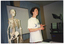 COL medium close up photo of a woman standing next to a plastic skeleton on a stand. She is wearing a t-shirt with the text and logo, 'Joint Action'. Beside her, there is a handwritten sheet of paper on a table (partially obscured). Behind her, there is a slide projector screen on a stand in front of two large curtained windows. To the right of the frame, there is a slide projector near a televison set on a stand. There is a note taped to the side of the television set which says, "VIDEO BOOKED OUT TO PHYSIO 29.5.96 TO 31.5.96”.