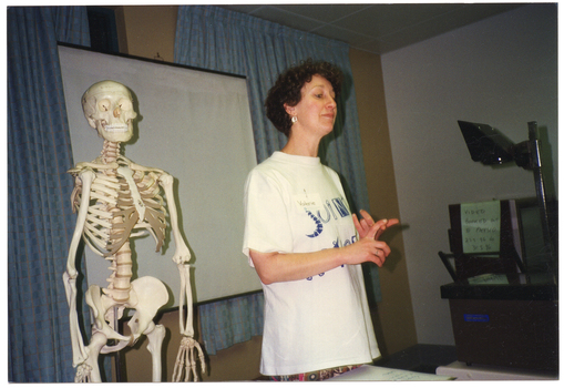 COL medium close up photo of a woman standing next to a plastic skeleton on a stand. She is wearing a t-shirt with the text and logo, 'Joint Action'. Beside her, there is a handwritten sheet of paper on a table (partially obscured). Behind her, there is a slide projector screen on a stand in front of two large curtained windows. To the right of the frame, there is a slide projector near a televison set on a stand. There is a note taped to the side of the television set which says, "VIDEO BOOKED OUT TO PHYSIO 29.5.96 TO 31.5.96”.