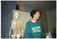 COL medium close up photo of a woman standing next to a plastic skeleton on a stand. She is wearing a jade green sweatshirt with the text and logo for the 'Move it or Lose it' program and the Arthritis Foundation of Victoria. She is holding a handwritten sheet of paper. There is a plastic skeleton on a vertical stand beside her, and a projector screen behind her. Behind the screen, there are two large curtained windows.