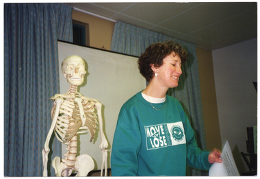 COL medium close up photo of a woman standing next to a plastic skeleton on a stand. She is wearing a jade green sweatshirt with the text and logo for the 'Move it or Lose it' program and the Arthritis Foundation of Victoria. She is holding a handwritten sheet of paper. There is a plastic skeleton on a vertical stand beside her, and a projector screen behind her. Behind the screen, there are two large curtained windows.