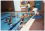 COL medium close up photo of several people at a swimming pool. A woman kneeling down on a thick blue mat next to the edge of the pool. She has one hand outstretched and the other hand placed high up on her side. There is a small group of people in the pool. One person is floating, supported by five other people. The remainder of the group is watching them. One person is standing at the far end of the pool deck, watching the group in the pool. Behind her, there are some shelves containing various floatation devices. There are two closed doors on either side of the shelves, and two yellow plastic chairs further along the wall beside one of the doors.