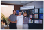 COL photo of three people standing in front of a multipanel display board. At the top of the display board, there is a sign with the text, "ARTHRITIS VICTORIA", and a logo. On the display board, there are various large photos, posters, books and brochures in clear perspex holders. Poster titles include "Stay on your feet", "Living a Healthy Life with Chronic Conditions", and "Exercise Beats Arthritis". Book titles include "The Arthritis Handbook" and "The Arthritis Helpbook". To the left of the display board, there is a brick wall with a large window. There is a large fern outside of the window.