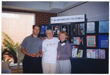 COL photo of three people standing in front of a multipanel display board. At the top of the display board, there is a sign with the text, "ARTHRITIS VICTORIA", and a logo. On the display board, there are various large photos, posters, books and brochures in clear perspex holders. Poster titles include "Stay on your feet", "Living a Healthy Life with Chronic Conditions", and "Exercise Beats Arthritis". Book titles include "The Arthritis Handbook" and "The Arthritis Helpbook". To the left of the display board, there is a brick wall with a large window. There is a large fern outside of the window.
