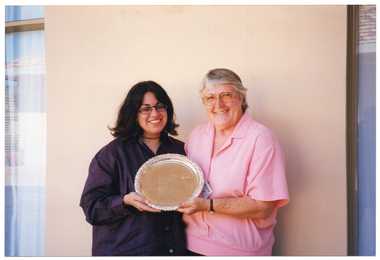 COL photo of two women, standing outside a building, are holding an engraved silver platter. There is a section of a wall behind them, with two curtained windows (partially obscured) on either side.