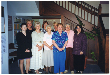 COL photo of a group of people posing for a photo inside a building. Behind them are a wall display of brochures, some directional signs indicating the location of the building's commercial tenants, and a wooden staircase leading to the next floor.