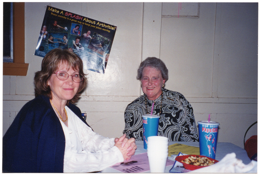 COL photo of two women seated at a table. On the table, there are two PhysiCal disposable milkshake cups, a small stack of polystyrene cups, a red bowl containing fruit and nut mix, and several printed leaflets in different colours. On the wall behind them, there is a poster titled "Make A SPLASH About Arthritis!", with the sub-title "Water exercise is a great way to keep your joints moving."