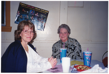 COL photo of two women seated at a table. On the table, there are two PhysiCal disposable milkshake cups, a small stack of polystyrene cups, a red bowl containing fruit and nut mix, and several printed leaflets in different colours. On the wall behind them, there is a poster titled "Make A SPLASH About Arthritis!", with the sub-title "Water exercise is a great way to keep your joints moving."