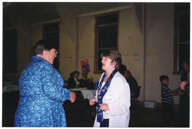COL photo of two women chatting and/or dancing in a large hall. In the background, there are small groups of people seated at tables. There are several tall windows along the wall near the tables.