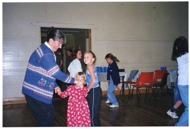 COL photo of some people dancing inside a large hall. A woman is dancing with a young child, and other children are dancing nearby. In the background, there are some empty tables and chairs, and some people are standing in an open double doorway.