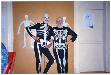 COL photo of two people wearing skeleton costumes. One person has his face covered in white tape or fabric to look like a skull. The other person has white fabric or bandages wrapped around her head and chin. Behind then, there are two paper or cardboard skeleton cut-outs stuck to two closed doors. On the wall, there is a piece of paper with a labelled drawing of a skeleton. To the left of the frame, the back of a person sitting on a chair is evident.