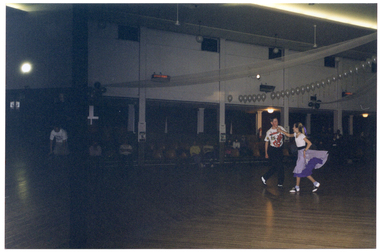 COL photo of a teenage dance couple performing a rock and roll dance in a large hall. He is wearing a colourful t-shirt and black trousers, while she is wearing a white t-shirt and a purple and mauve circle skirt. Several people are watching the performance while seated in rows of seats. There is a row of white balloons decorating the hall in the ceiling space.