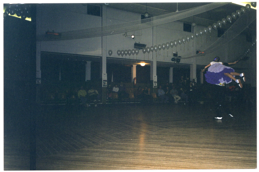 COL photo of a teenage dance couple performing a rock and roll dance in a large hall. He is wearing a colourful t-shirt and black trousers, while she is wearing a white t-shirt and a purple and mauve circle skirt. The male dancer is lifting the female up in the air. Several people are watching the performance while seated in rows of seats. There are several rows of white balloons decorating the hall in the ceiling space.