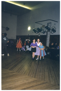 COL photo of three young dancers performing a promenade dance step in a large hall. They are each wearing a full circle skirt with a plain top. To the left of the frame, there is another person, dressed in jeans and a t-shirt, dancing.  In the background, there are several people either standing or seated. Behind them, there is a large cluster of white and teal green balloons suspended from a small fenced platform.
