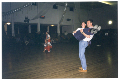 COL photo of two couples dancing in a large hall. One couple has the man crouching down while the woman is standing behind him with one hand on his head. They are wearing matching costumes consisting of black and white checked shirts with red trousers. The other couple has the man carrying the woman horizontally in his arms. They are dressed in contemporary jeans and shirts. Several people are watching the performance while seated in rows of seats on the periphery. There are rows of white balloons decorating the hall in the ceiling space.
