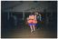 COL photo of several couples doing rock and roll dancing on the dancefloor in a large hall. One couple is dancing in the foreground. The woman is wearing an orange dress with a  purple petticoat and  the man is wearing a silver sequined jacket with black pants and a black Elvis Presley t-shirt. Several people are watching the performance while seated in rows of seats on the periphery. There are strings and clusters of white balloons decorating the hall in the ceiling space.
