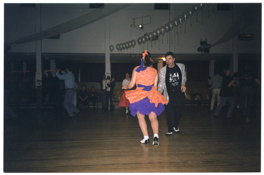 COL photo of several couples doing rock and roll dancing on the dancefloor in a large hall. One couple is dancing in the foreground. The woman is wearing an orange dress with a  purple petticoat and  the man is wearing a silver sequined jacket with black pants and a black Elvis Presley t-shirt. Several people are watching the performance while seated in rows of seats on the periphery. There are strings and clusters of white balloons decorating the hall in the ceiling space.