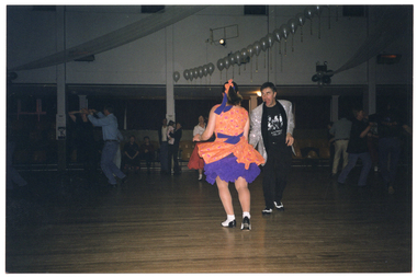 COL photo of several couples doing rock and roll dancing on the dancefloor in a large hall. One couple is dancing in the foreground. The woman is wearing an orange dress with a  purple petticoat and  the man is wearing a silver sequined jacket with black pants and a black Elvis Presley t-shirt. Several people are watching the performance while seated in rows of seats on the periphery. There are strings and clusters of white balloons decorating the hall in the ceiling space.