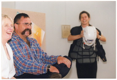 COL photo of three people indoors, one standing and two seated. The woman standing is holding up a protective sports helmet with both hands. She is looking towards the two seated people, who are looking off camera. All three people are laughing.