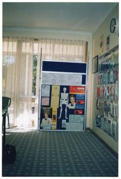 COL photo of a large, freestanding display board in front of a curtained window. Beside the display board, on a perpendicular wall, there is a display of numerous brochures inside plastic sleeves affixed to wall. There are also a few signs or posters, a framed item, and a clock on the wall.