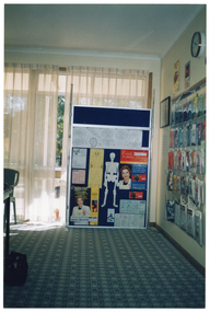 COL photo of a large, freestanding display board in front of a curtained window. Beside the display board, on a perpendicular wall, there is a display of numerous brochures inside plastic sleeves affixed to wall. There are also a few signs or posters, a framed item, and a clock on the wall.