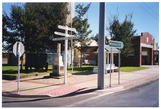 COL photo of some directional road signs, a cenotaph, and a wooden bench with an overhead shelter on a paved area next to a building. In the foreground, one directional sign indicates 'CALDER HIGHWAY' and directions to various locations nearby, such as: 77 WYCHEPROOF; OUYEN (partially obscured); 33 LASCELLES; and SWAN HILL 77. A larger green directional sign indicates Route A79 to the town of Mildura.
