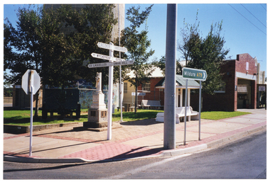 COL photo of some directional road signs, a cenotaph, and a wooden bench with an overhead shelter on a paved area next to a building. In the foreground, one directional sign indicates 'CALDER HIGHWAY' and directions to various locations nearby, such as: 77 WYCHEPROOF; OUYEN (partially obscured); 33 LASCELLES; and SWAN HILL 77. A larger green directional sign indicates Route A79 to the town of Mildura.