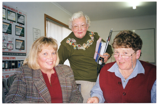 COL photo of three women posing for a photo at a table or desk. Two of the women are seated while the third woman is standing between them. The woman standing is holding a stack of brochures and leaflets and a clipboard. On the wall behind them is a large display board with headings such as "AND PEN NEEDLES..." and "TESTING STRIPS". In the background, there is a window with vertical blinds.