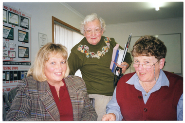 COL photo of three women posing for a photo at a table or desk. Two of the women are seated while the third woman is standing between them. The woman standing is holding a stack of brochures and leaflets and a clipboard. On the wall behind them is a large display board with headings such as "AND PEN NEEDLES..." and "TESTING STRIPS". In the background, there is a window with vertical blinds.