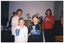 COL photo of two girls and two women standing indoors. The two girls and one of the women are each holding a disposable milkshake container in one hand. There two other people in the background, (partially obscured) standing behind a table with an electric milkshake maker appliance, bottles of milk and flavoured syrup. On the wall, there are two posters with a skeleton graphic and the text, "NATIONAL HEALTHY BONES WEEK YOUR BONES YOUR LIFE".